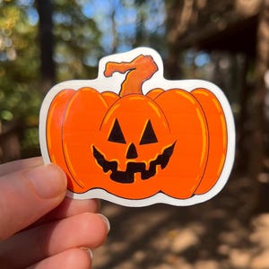 May include: A Halloween-themed sticker featuring a smiling jack-o'-lantern. The pumpkin is orange with a black face and a brown stem. The sticker has a white border and is held against a blurred outdoor background.
