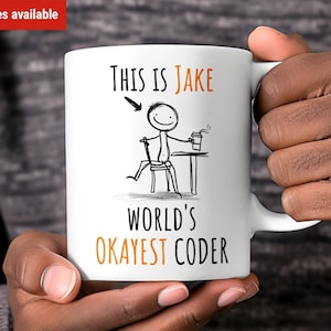 May include: White ceramic mug featuring a black stick figure illustration of a person seated at a desk with a cup of coffee. The text "This is Jake World's Okayest Coder" is printed on the mug in black and orange.