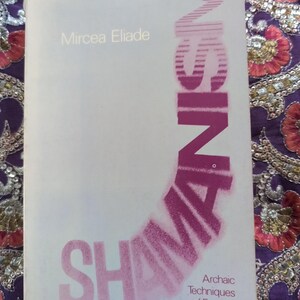 May include: A white book cover with the title "SHAMANISM" in pink lettering. The book is by Mircea Eliade and is about archaic techniques of ecstasy. The book is part of the Bollingen Series and was published by Princeton University Press for $4.95.