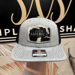 May include: A gray and black trucker hat with a silver patch that says "Gulf of America est. 2025". The hat has a black mesh back and a Richardson 112 adjustable strap.