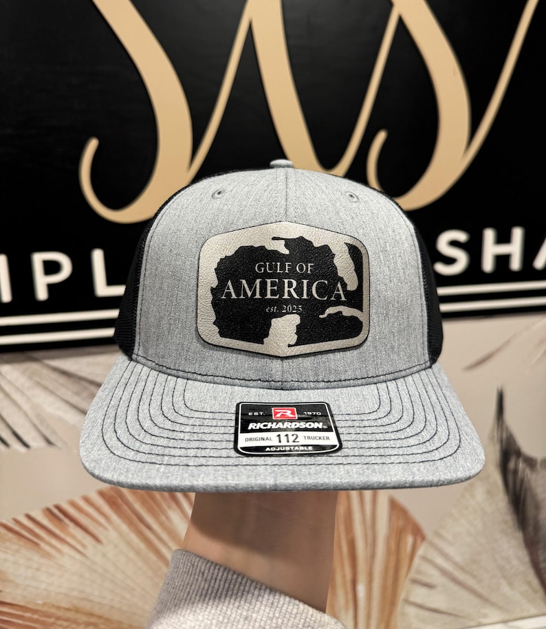 K&ouml;nnte beinhalten: Eine graue und schwarze Trucker-Kappe mit einem silbernen Patch, auf dem "Gulf of America est. 2025" steht. Die Kappe hat ein schwarzes Netzr&uuml;cken und einen verstellbaren Richardson 112-Riemen.