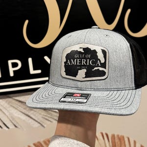 K&ouml;nnte beinhalten: Eine graue und schwarze Trucker-Kappe mit einem schwarzen Patch, auf dem "Gulf of America est. 2025" steht.