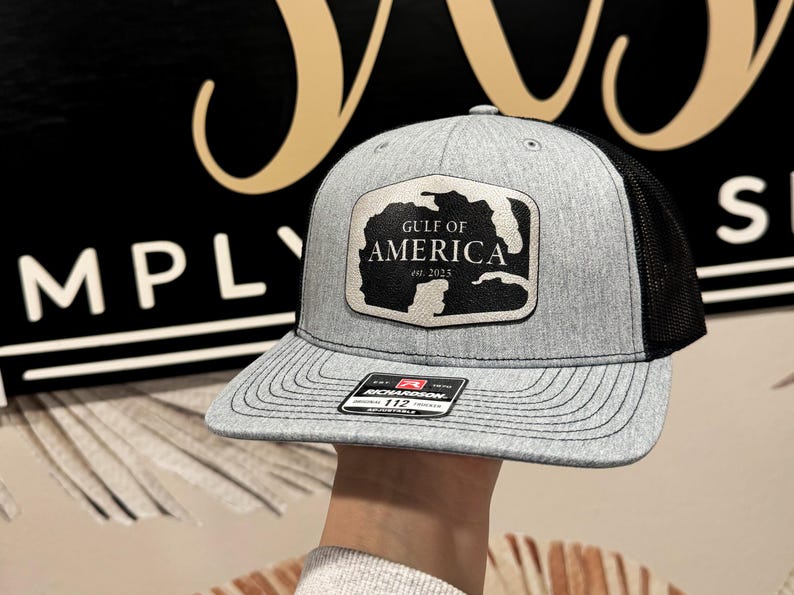 K&ouml;nnte beinhalten: Eine graue und schwarze Trucker-Kappe mit einem schwarzen Patch, auf dem "Gulf of America est. 2025" steht.
