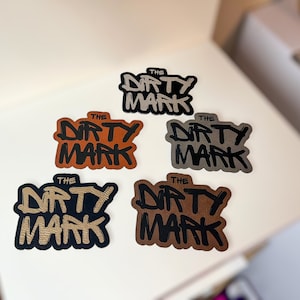 K&ouml;nnte beinhalten: F&uuml;nf verschiedene farbige Patches mit dem Text "THE DIRTY MARK" in schwarzer Schrift. Die Patches sind aus Leder und haben einen Used-Look.