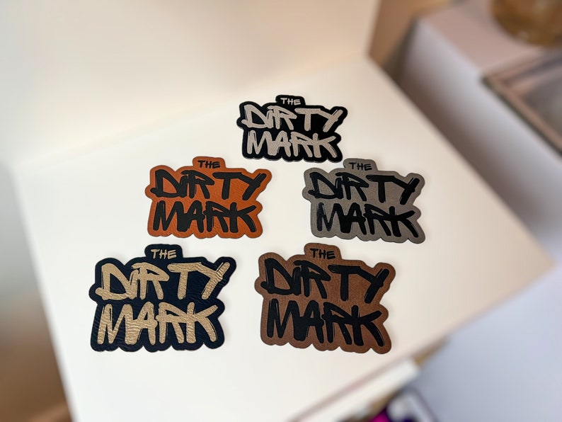 K&ouml;nnte beinhalten: F&uuml;nf verschiedene farbige Patches mit dem Text "THE DIRTY MARK" in schwarzer Schrift. Die Patches sind aus Leder und haben einen Used-Look.