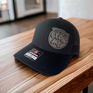 May include: Black trucker hat with a black mesh back. The hat has a gray patch with the text "Why don't you blow me" in a bubble font.