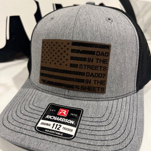 May include: A gray and black trucker hat with a brown leather patch featuring a distressed American flag and the text "DAD IN THE STREETS DADDY IN THE SHEETS". The hat has a black mesh back and a Richardson 112 adjustable strap.