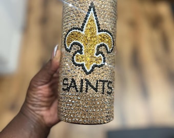 May include: A gold-colored, bedazzled tumbler with a fleur-de-lis logo in black, gold, and white. The word "SAINTS" is written in black below the logo. The tumbler is covered in small, sparkling rhinestones.