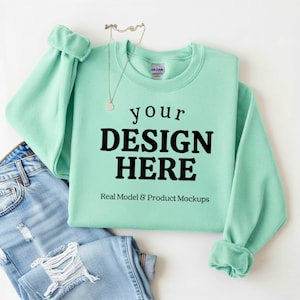 May include: A mint green sweatshirt with the text "your DESIGN HERE" in bold black letters. The sweatshirt is paired with light blue ripped jeans and a delicate necklace. The words "Real Model & Product Mockups" are below the main text.