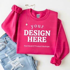 May include: A magenta sweatshirt with the text "your DESIGN HERE" in white, with a pair of light blue jeans. A silver necklace with a pendant is on top of the sweatshirt. The sweatshirt has a crew neck and ribbed cuffs.