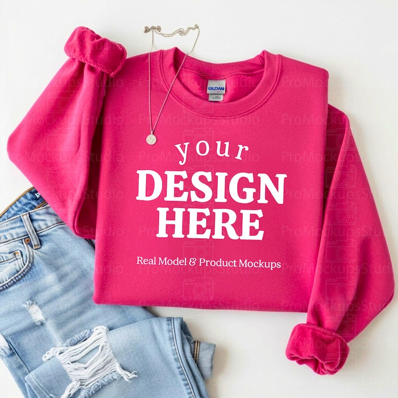 May include: A magenta sweatshirt with the text "your DESIGN HERE" in white, with a pair of light blue jeans. A silver necklace with a pendant is on top of the sweatshirt. The sweatshirt has a crew neck and ribbed cuffs.