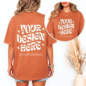 May include: A rust-colored t-shirt with the text "YOUR DESIGN HERE" in a white, retro font. The shirt has short sleeves and a relaxed fit. The text "ADD YOUR DESIGN WITH PHOTOSHOP, CANVA OR OTHER EDITING PROGRAM" is below the main text.
