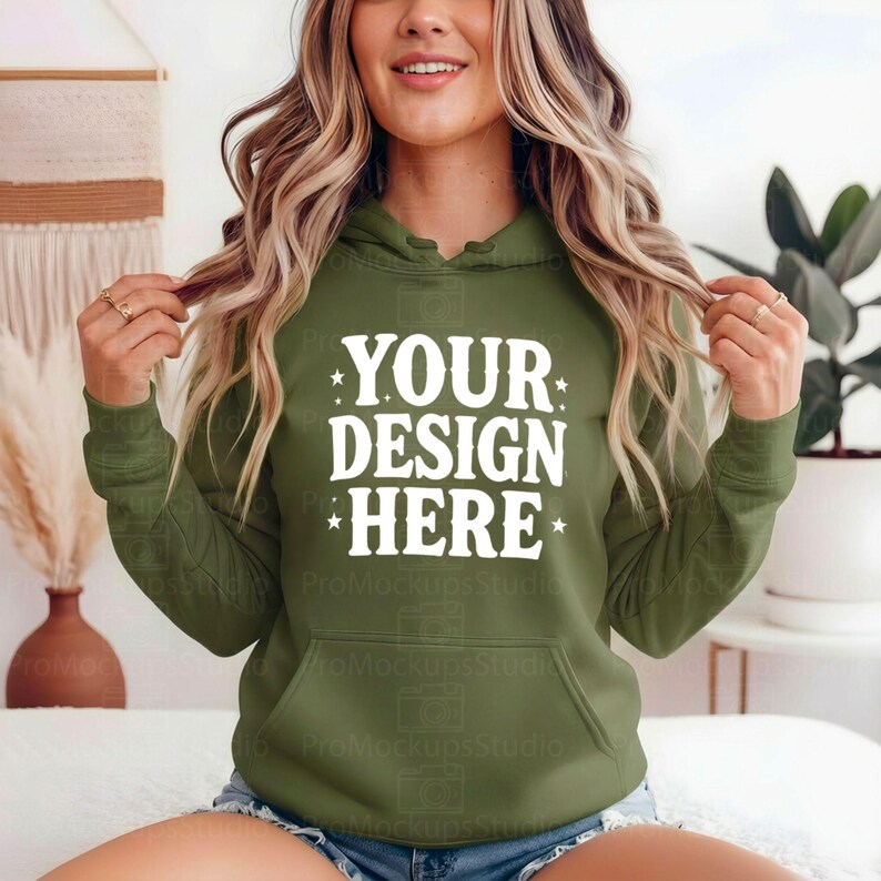 K&ouml;nnte beinhalten: Ein olivgr&uuml;ner Kapuzenpullover mit dem Text "YOUR DESIGN HERE" in Wei&szlig;. Der Kapuzenpullover hat eine Vordertasche und eine Kapuze. Das Model tr&auml;gt Jeansshorts. Der Hintergrund umfasst einen dekorativen Wandbehang und eine Pflanze.