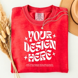 May include: A red t-shirt with the text "YOUR DESIGN HERE" in white, with star accents. A silver necklace with a star pendant is draped over the shirt. The shirt has a Comfort Colors tag. A tan hat is in the background.