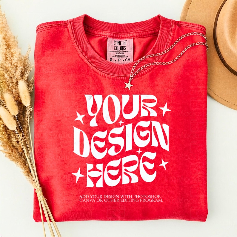 May include: A red t-shirt with the text "YOUR DESIGN HERE" in white, with star accents. A silver necklace with a star pendant is draped over the shirt. The shirt has a Comfort Colors tag. A tan hat is in the background.