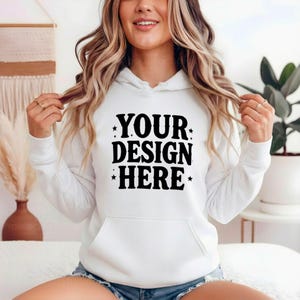K&ouml;nnte beinhalten: Ein wei&szlig;er Kapuzenpullover mit einer Vordertasche und einem Kordelzug. Der Text "YOUR DESIGN HERE" ist in Schwarz auf der Vorderseite aufgedruckt. Dieses l&auml;ssige Kleidungsst&uuml;ck eignet sich f&uuml;r verschiedene Designanwendungen.