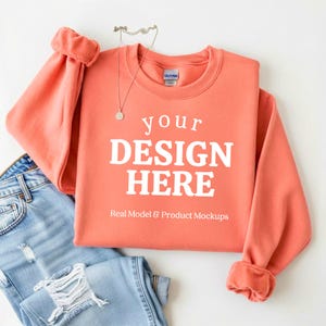 May include: A coral-colored sweatshirt with the text "your DESIGN HERE" in white, with a pair of light blue jeans. A silver necklace with a small pendant is draped over the sweatshirt. The sweatshirt has cuffed sleeves.