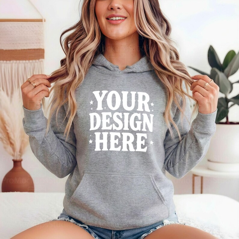 K&ouml;nnte beinhalten: Ein melierter grauer Kapuzenpullover mit einer Vordertasche und einer Kapuze mit Kordelzug. Der Text "YOUR DESIGN HERE" ist wei&szlig; auf der Vorderseite aufgedruckt. Der Kapuzenpullover wird von einer Person getragen.