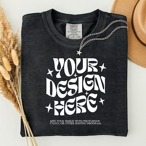 May include: Black t-shirt with the text "YOUR DESIGN HERE" in white, with star accents. A silver necklace with a star pendant and a brown hat are also visible. The shirt has a Comfort Colors tag.