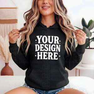 K&ouml;nnte beinhalten: Schwarzer Kapuzenpullover mit dem wei&szlig;en Text "YOUR DESIGN HERE". Der Pullover hat eine Vordertasche und eine Kapuze mit Kordelzug. Das Design ist mittig auf der Brust platziert.