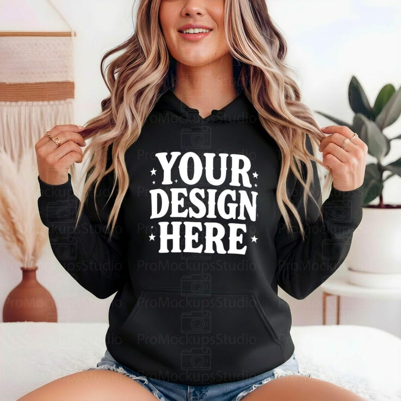 K&ouml;nnte beinhalten: Schwarzer Kapuzenpullover mit dem wei&szlig;en Text "YOUR DESIGN HERE". Der Pullover hat eine Vordertasche und eine Kapuze mit Kordelzug. Das Design ist mittig auf der Brust platziert.