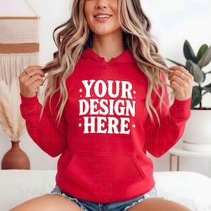K&ouml;nnte beinhalten: Ein roter Kapuzenpullover mit einer Vordertasche und einer Kapuze mit Kordelzug. Der Text "YOUR DESIGN HERE" ist in Wei&szlig; auf die Brust gedruckt. Der Kapuzenpullover wird von einer Person getragen.