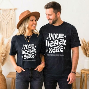 Op de afbeelding: Twee mensen dragen zwarte t-shirts met de tekst "YOUR DESIGN HERE" in het wit, met steraccenten. De shirts hebben korte mouwen. De vrouw draagt een bruine hoed en de man heeft een baard.