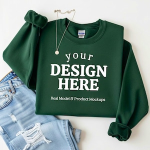 May include: A dark green sweatshirt with the text "your DESIGN HERE" in white, with a silver necklace and a pair of blue jeans. The sweatshirt has cuffed sleeves and is a mockup for design placement.