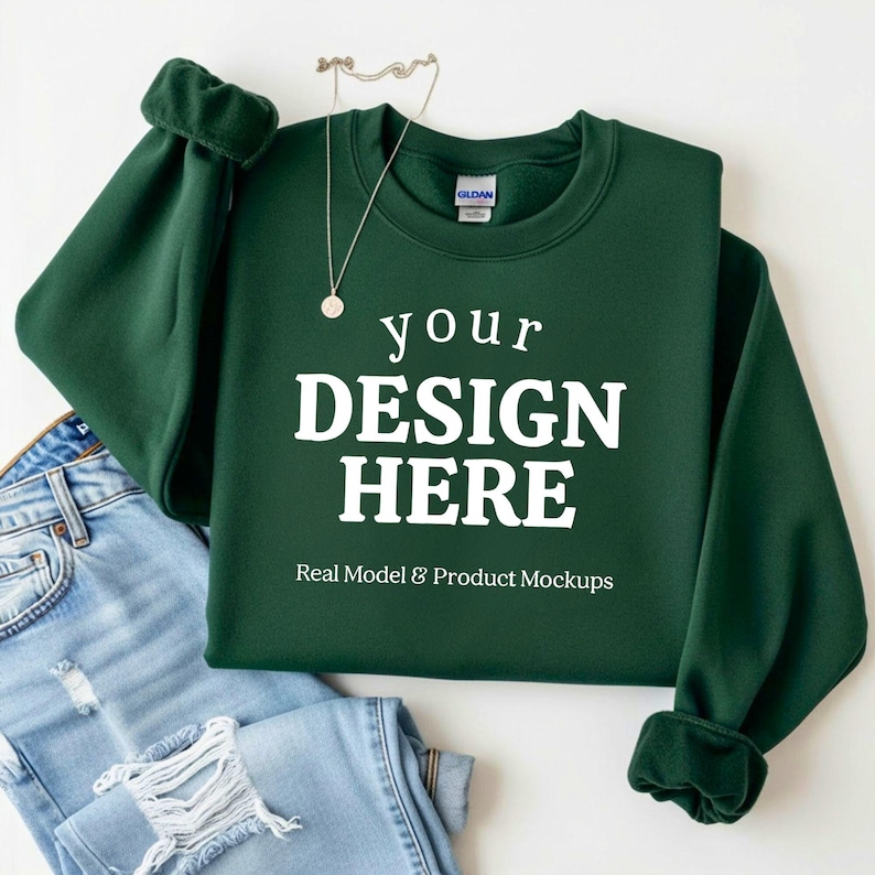 May include: A dark green sweatshirt with the text "your DESIGN HERE" in white, with a silver necklace and a pair of blue jeans. The sweatshirt has cuffed sleeves and is a mockup for design placement.