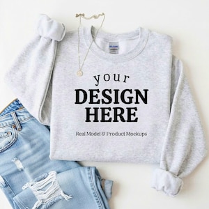 May include: A light gray sweatshirt with the text "your DESIGN HERE" in black, with the words "Real Model & Product Mockups" below. The sweatshirt is paired with light blue denim jeans and a gold necklace.