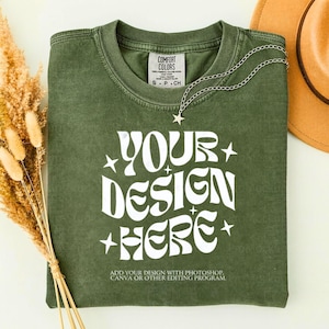 May include: Olive green t-shirt with the text "YOUR DESIGN HERE" in white, with star accents. A silver necklace with a star pendant is draped on the shirt. The shirt has a small tag that says "COMFORT COLORS".