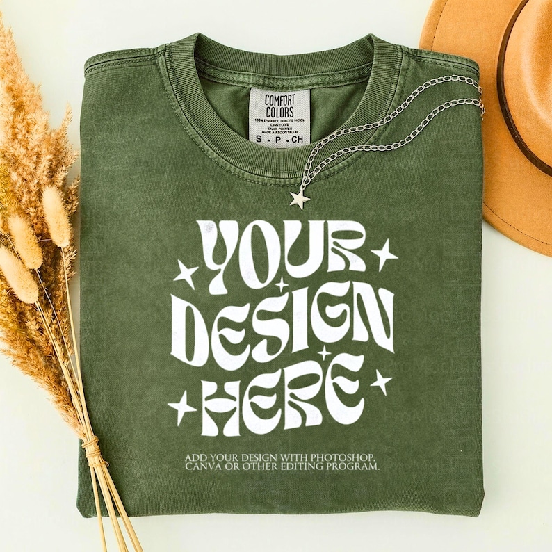May include: Olive green t-shirt with the text "YOUR DESIGN HERE" in white, with star accents. A silver necklace with a star pendant is draped on the shirt. The shirt has a small tag that says "COMFORT COLORS".