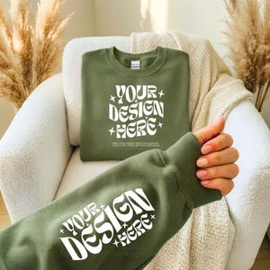Peut inclure: Un sweat-shirt vert olive avec le texte "YOUR DESIGN HERE" en blanc, dans une police rétro. Le sweat-shirt est plié sur une chaise blanche avec une couverture beige. La manche du sweat-shirt est visible, avec le même texte.