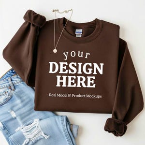 May include: A brown sweatshirt with the text "your DESIGN HERE" in white. The sweatshirt is displayed with a pair of blue jeans and a silver necklace. The words "Real Model & Product Mockups" are also visible.