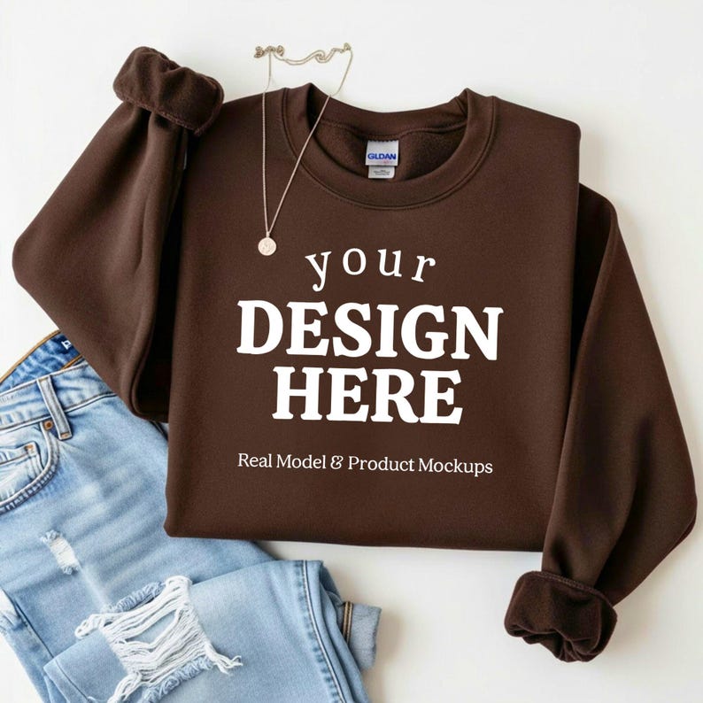 May include: A brown sweatshirt with the text "your DESIGN HERE" in white. The sweatshirt is displayed with a pair of blue jeans and a silver necklace. The words "Real Model & Product Mockups" are also visible.