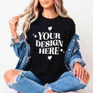 Puede incluir: Camiseta negra con el texto "YOUR DESIGN HERE" en blanco, adornada con corazones y estrellas. La modelo lleva una chaqueta vaquera y unos vaqueros azules rotos.
