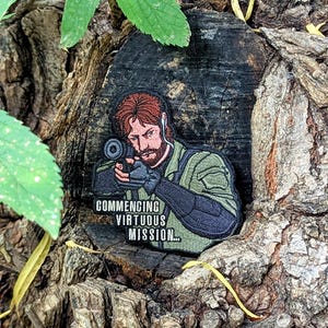 May include: Embroidered patch featuring a man aiming a weapon, with red hair and a beard. The patch is green, black, and tan, with the text "COMMENCING VIRTUOUS MISSION..." in white. The patch is set against a natural wood background.