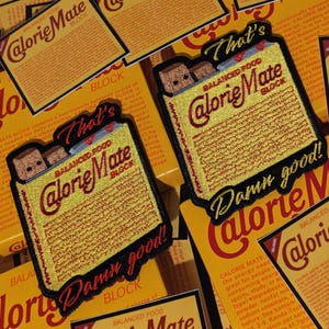 Op de afbeelding: Geborduurde patch met een geel Calorie Mate Block-ontwerp met de tekst "That's" en "Damn good!" in zwart en rood. De patch toont een afbeelding van het voedselproduct en is omgeven door vergelijkbare verpakkingen.
