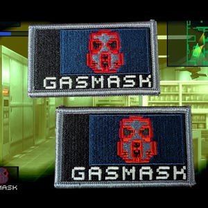 May include: Two rectangular embroidered patches featuring a pixelated red gas mask icon centered on a dark blue background, with the word GASMASK stitched in white block letters below. The patches have a black side panel and a grey border.