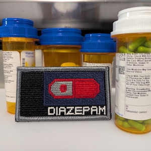 Può includere: Immagine di tre flaconi di medicinali con coperchi blu e uno con coperchio bianco, pieni di pillole. Una toppa con la scritta "DIAZEPAM" e un disegno di pillola è davanti ai flaconi. Le pillole sono verdi.