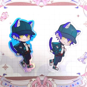 May include: Two holographic anime-style character stickers. Each sticker features a chibi character with blue hair, yellow eyes, and cat ears, wearing a dark green cap and jacket. The character has a blue tail and is standing on a clear base.