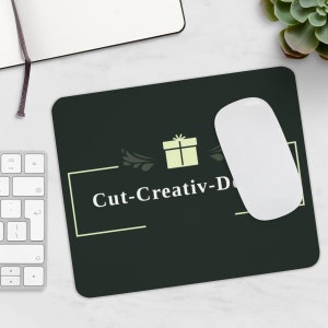 Könnte beinhalten: Ein schwarzes rechteckiges Mauspad mit dem Text "Cut-Creativ-Design" in Weiß. Eine weiße Computermaus liegt auf dem Mauspad. Das Mauspad zeigt auch ein hellgrünes Geschenkbox-Symbol und dekorative Verzierungen.