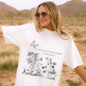 Könnte beinhalten: Weißes T-Shirt mit einer schwarz-weißen Illustration eines Schmetterlings, Blumen und Erdbeeren. Das Shirt trägt ein Zitat aus J.R.R. Tolkiens Der Herr der Ringe: "Es gibt Gutes in dieser Welt, Mr. Frodo, und es ist das Kämpfen wert."