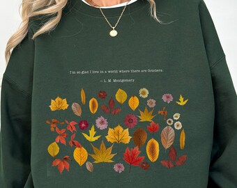 May include: Dark green sweatshirt with a white collar featuring a colorful leaf and flower design. The text reads, "I'm so glad I live in a world where there are Octobers." - L.M. Montgomery.