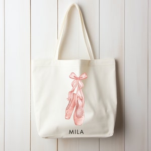 May include: A white canvas tote bag featuring a pink watercolour illustration of ballet shoes and a bow. The bag is personalised with the name "MILA" printed in black text.