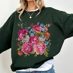Peut inclure: Un sweat-shirt vert foncé avec un imprimé floral de fleurs roses, rouges et blanches. Les fleurs sont disposées en bouquet et entourées de feuilles vertes.