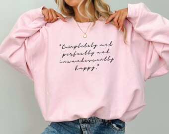 Puede incluir: Una sudadera rosa con el texto "Completamente y perfectamente e incandescentemente feliz." impreso en la parte delantera.