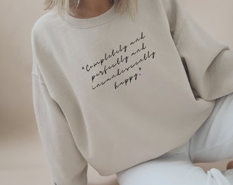 Puede incluir: Una sudadera beige con el texto "Completely and perfectly and incandescently happy." impreso en la parte delantera.