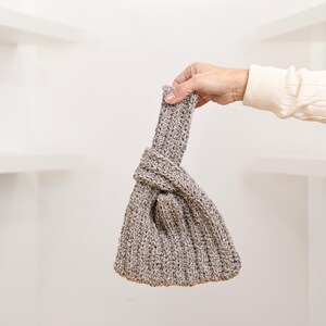May include: A gray, hand-knitted bag with a knotted design. The bag is held up by a hand, showcasing the texture and shape of the knitted material. The bag's color is a mix of gray and beige.