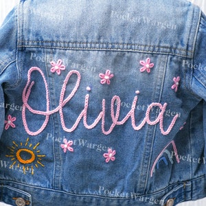 Peut inclure: Une veste en jean bleue avec une broderie rose du nom "Olivia" et de petites fleurs roses. La veste a également un soleil jaune et un arc-en-ciel brodés au dos.